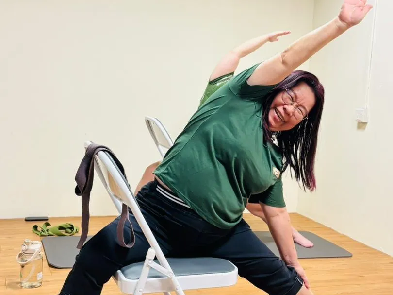 A smiling woman seated on a folding chair with her feet on yoga blocks, reaching one arm overhead in a side stretch, during a chair yoga class at Persistence Chiropractic Care in Cheras, Kuala Lumpur