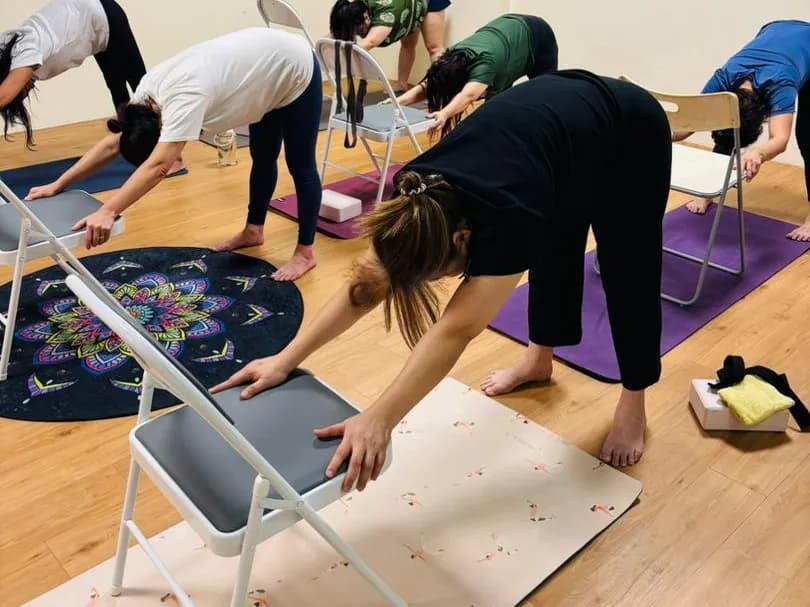 Students folding forward from standing with their hands resting on the seats of folding chairs during a yoga class at Persistence Chiropractic Care in Cheras, Kuala Lumpur