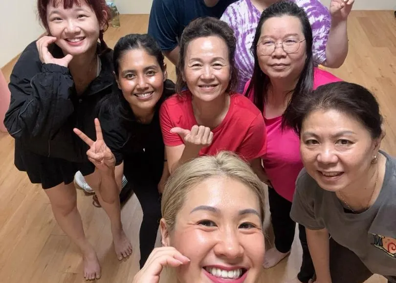 Seven smiling students posing for a group photo after a yoga class at Persistence Chiropractic Care in Cheras, Kuala Lumpur, with a silver yoga figurine and shelves of props behind them
