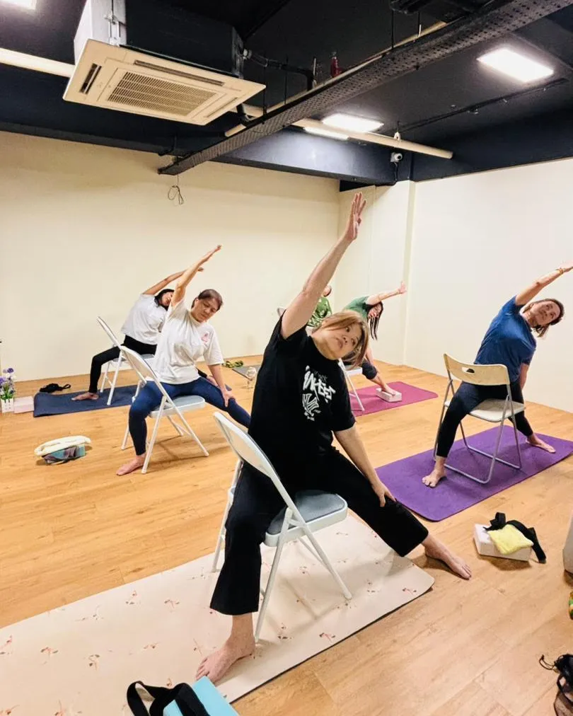 在 Cheras, Kuala Lumpur 的 Persistence Chiropractic Care 椅子瑜伽课上,五名学员坐在瑜伽垫上的折叠椅,各自单手举过头顶做侧伸展