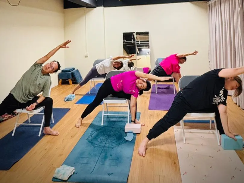 Students in a wide standing lunge with one arm reaching overhead, each beside a folding chair, during a yoga class at Persistence Chiropractic Care in Cheras, Kuala Lumpur